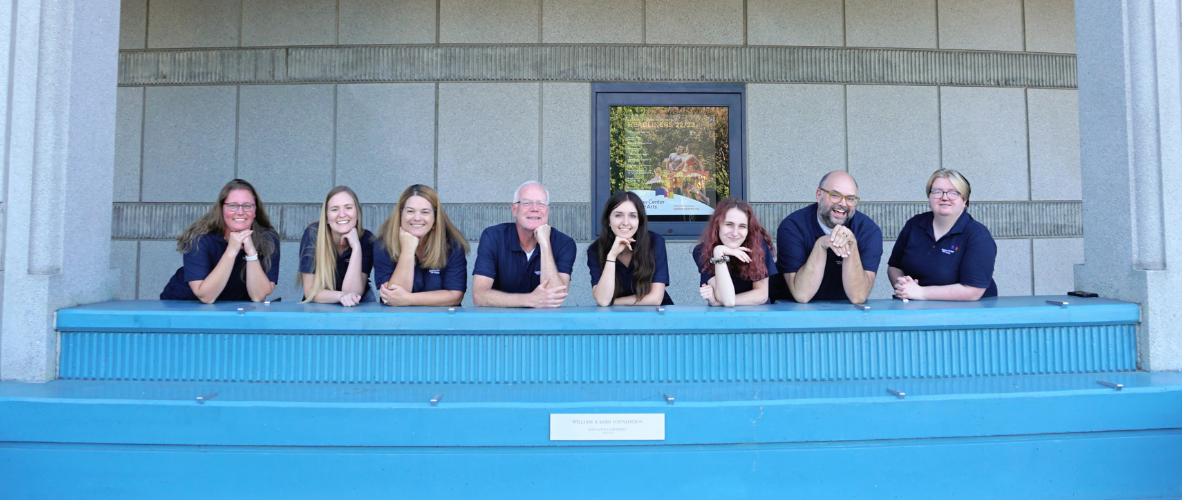 Group of 8 people pose behind a blue bench and against a building exterior